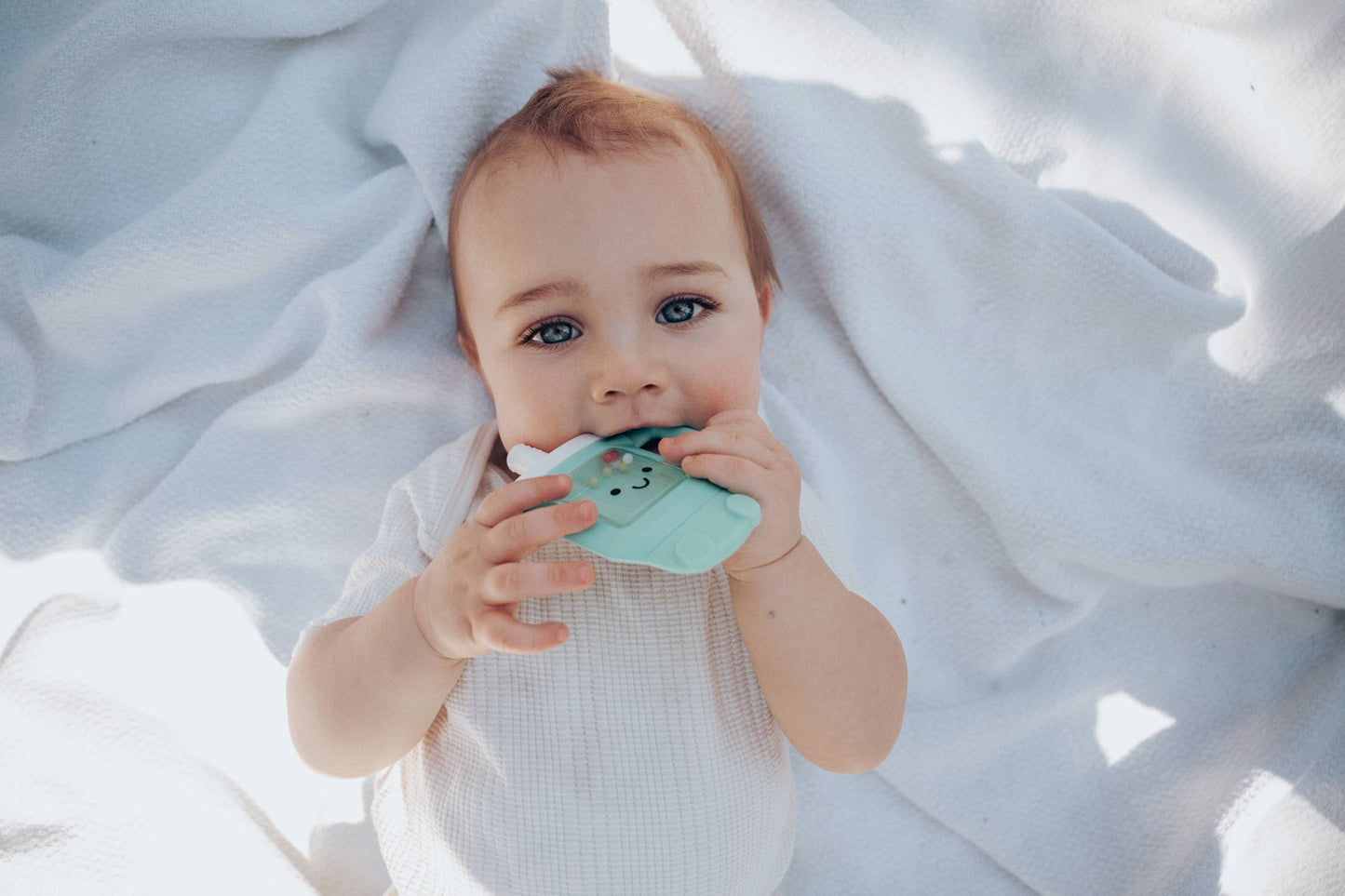 Baby holding a green teething toy on a white blanket