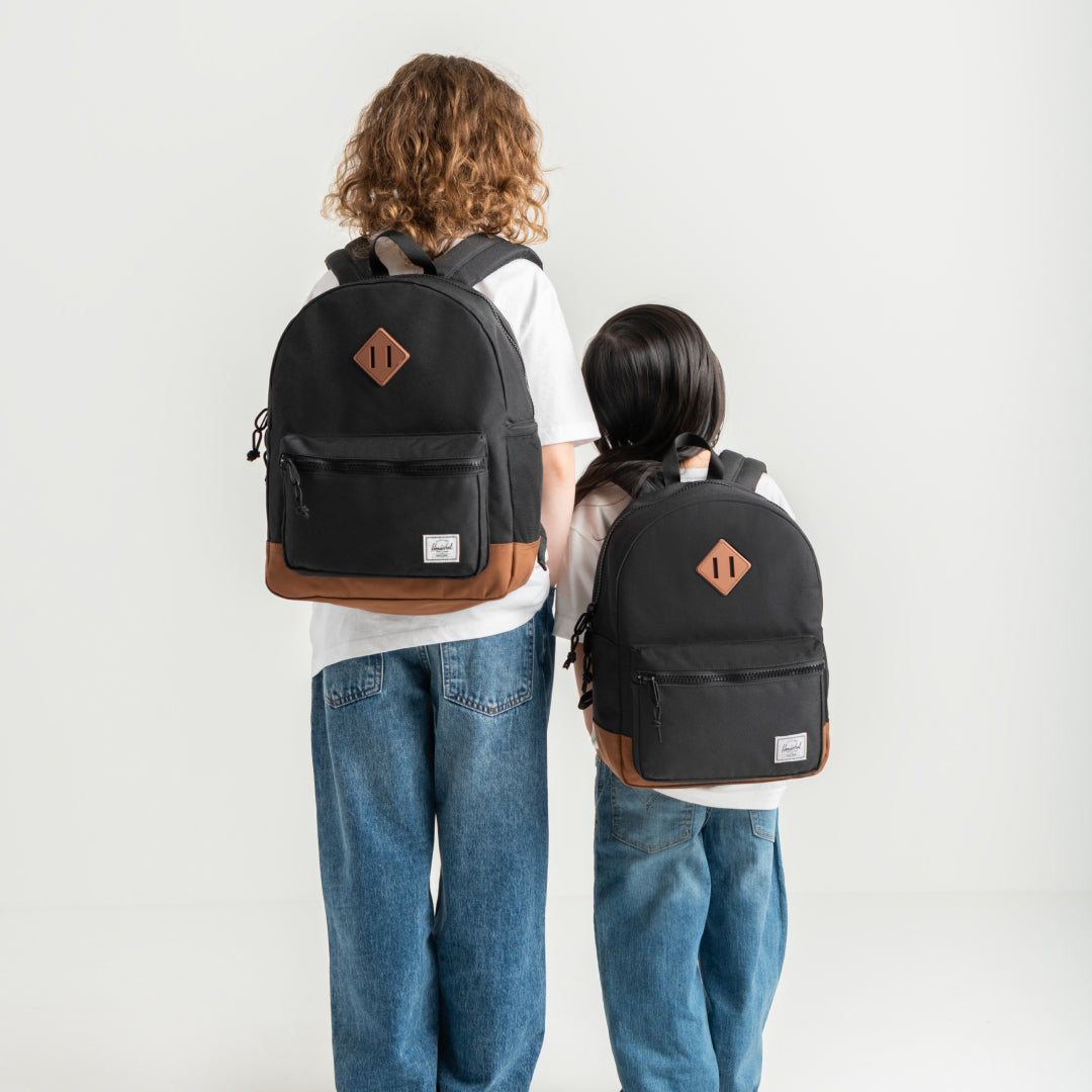Two children shown from behind wearing matching black backpacks with brown leather-like trim and a small diamond-shaped logo patch on the back, against a plain light gray studio background.